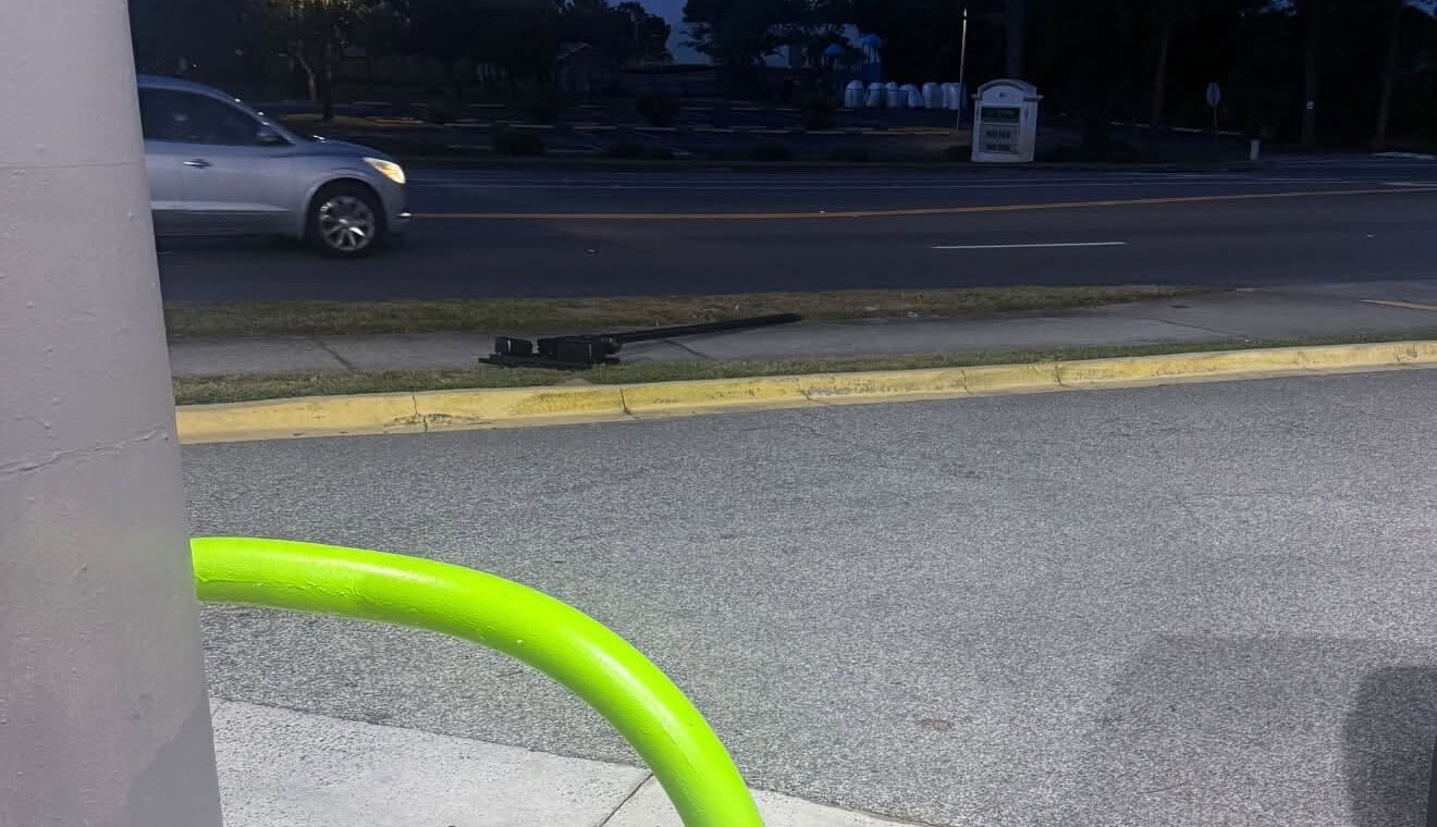 Dusk at Main Street and Airport Road in Destin. Across the road, a Flock license plate reader camera lies on the grass strip beside the curb.